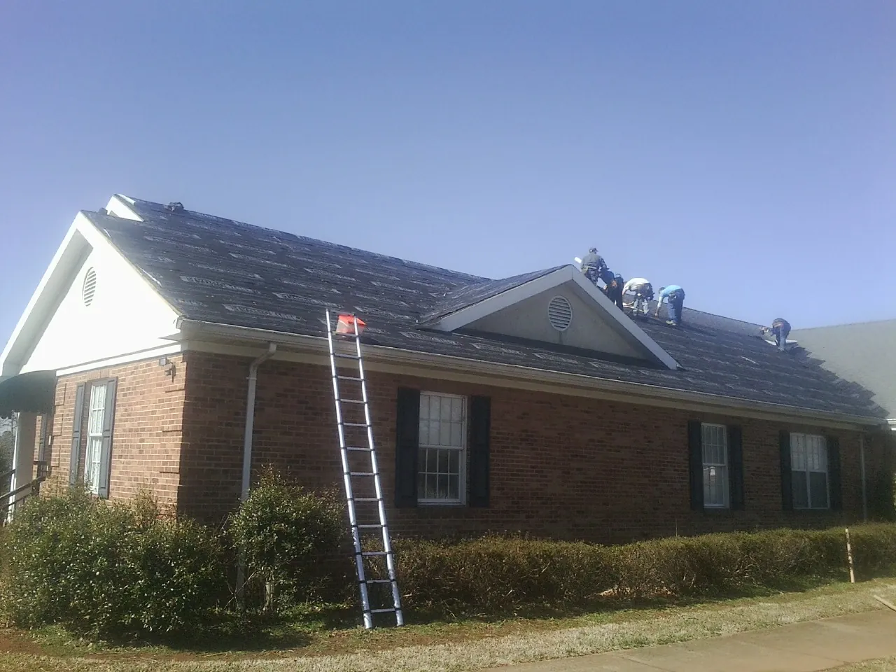 Crew tearing shingles off a brick ranch roof with a ladder set — Emergency Roof Repair work by our crew in Jeanerette