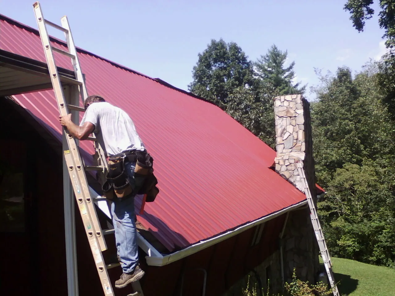 Roofer on a ladder beside a red metal roof and stone chimney — Soffit & Fascia Repair by Roofing Brothers in Jeanerette